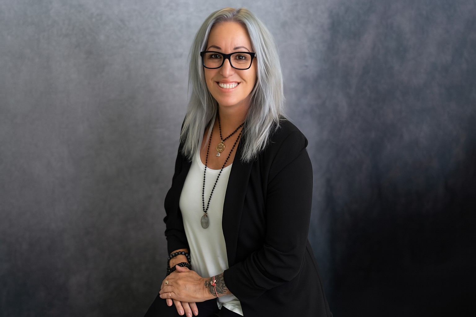 Woman with long silver hair wearing glasses, a black blazer, and necklaces sitting in front of a gray studio background.