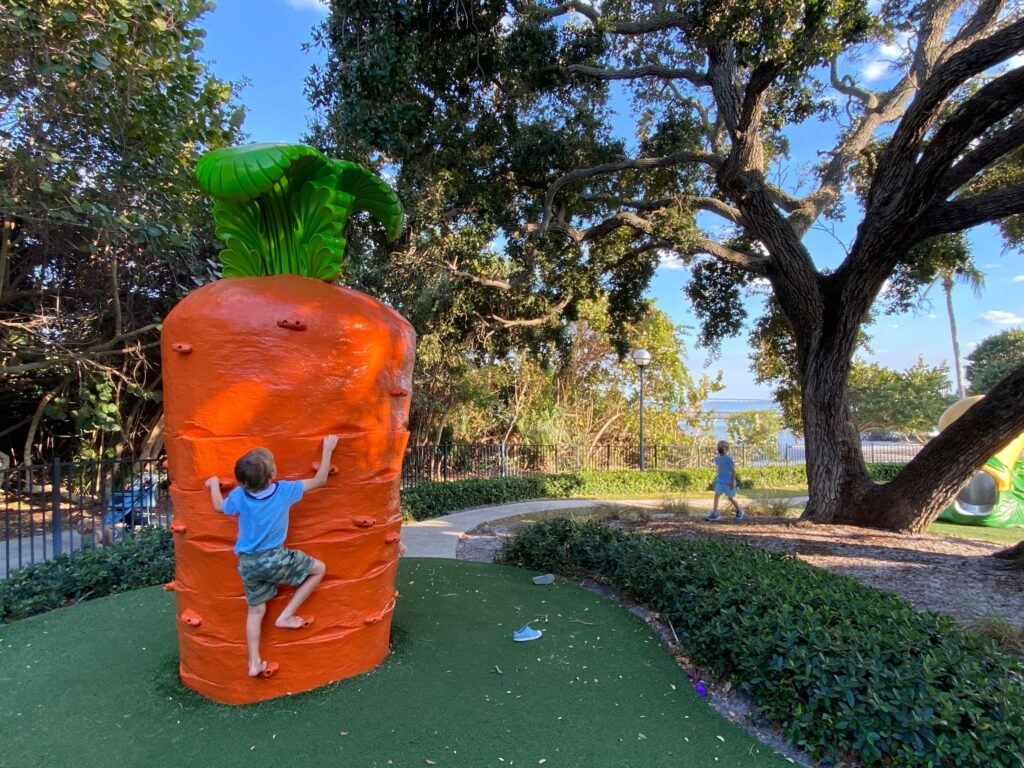 Child climbing a large orange carrot-shaped play structure beneath a large tree, with another child walking on a nearby path in a park.