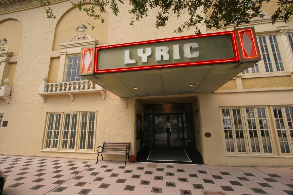 Lyric Theatre entrance featuring a vintage marquee sign with neon lighting above double glass doors and a bench on a tiled sidewalk.