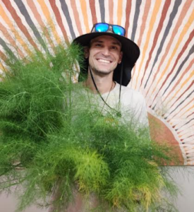 Smiling man in a wide sun hat and sunglasses holding a bunch of green, feathery plants, standing in front of a tapestry with radiating stripes.