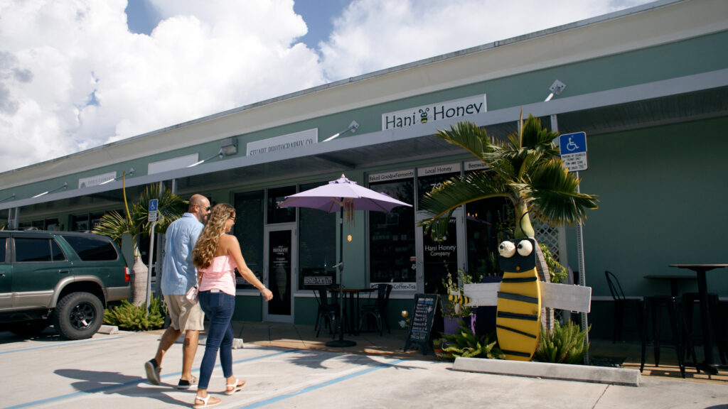 Couple walking in front of Hani Honey shop with outdoor seating, palm plants, and a large yellow bee sculpture by the entrance.