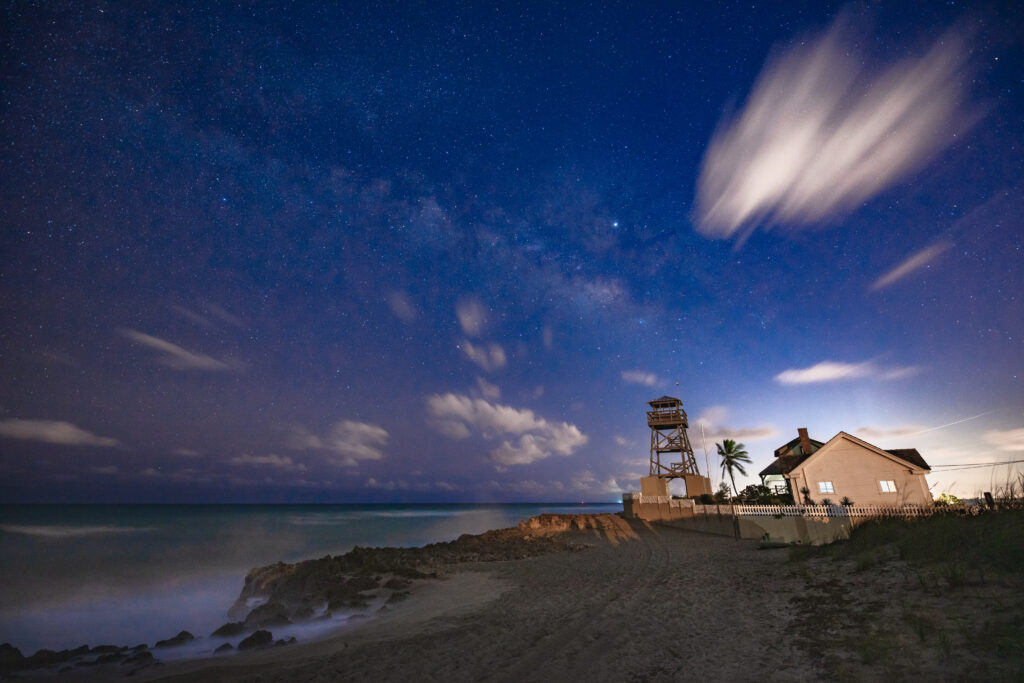 Nighttime beach scene with rocky shore, historic keeper's house, coastal watchtower, and a starry sky filled with wispy clouds.