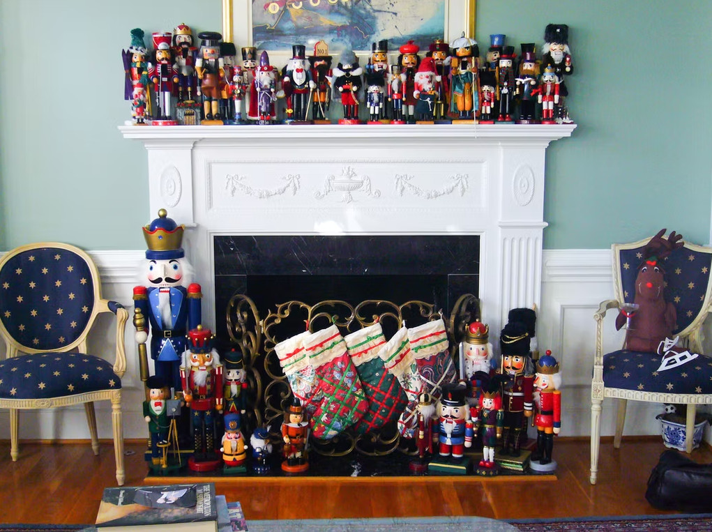 Collection of nutcracker and holiday figures arranged on and in front of a white fireplace mantel with three Christmas stockings