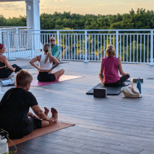 Small group of people sit on yoga mats for an outdoor class on a rooftop with a railing, facing trees and sunset light.
