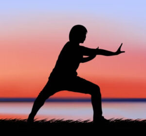 Silhouette of a person in a warrior yoga pose on grass by calm water at sunset, arms extended against a peaceful red-blue sky.