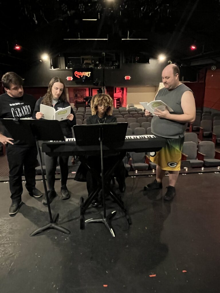 Four people gather around a keyboard on a theater stage, reading from music books, with empty audience seats behind them.