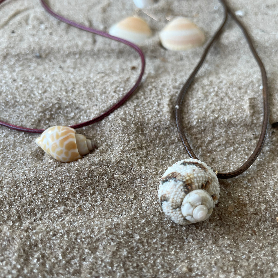 Two shell necklaces resting on sand with additional shells in the background.