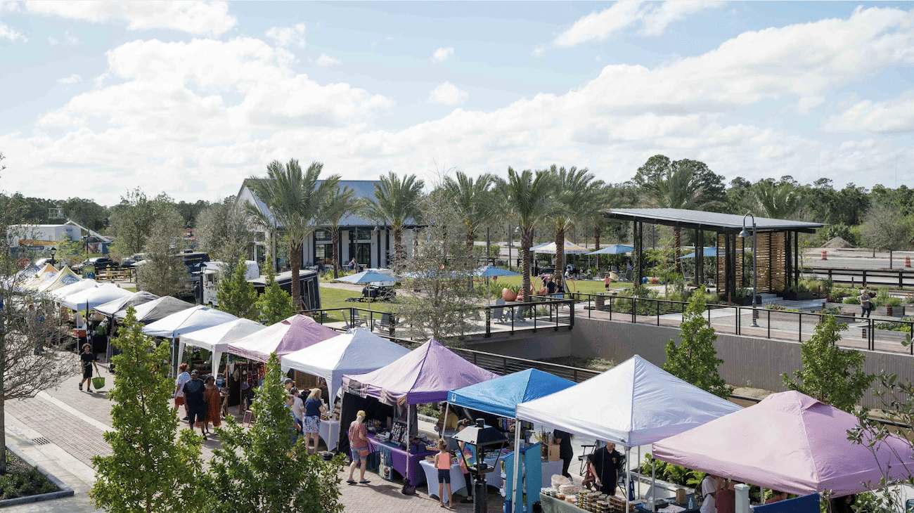 Outdoor market scene with colorful tents and palm trees, people browsing vendors under a sunny sky.