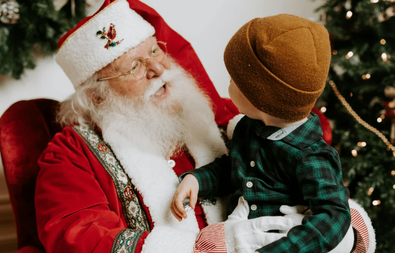 Child sitting on Santa’s lap during a holiday visit, with decorated tree in the background.