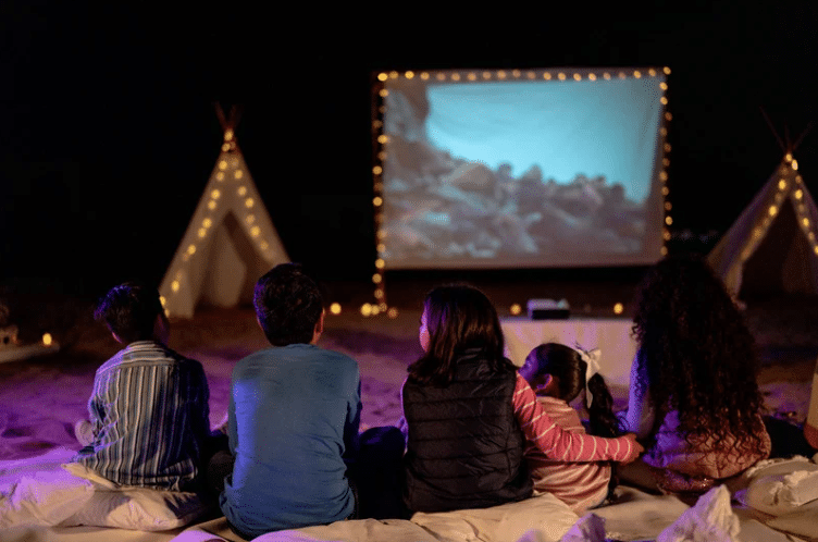 Five children sitting on blankets outdoors at night, watching a movie projected on a screen with illuminated tents nearby.
