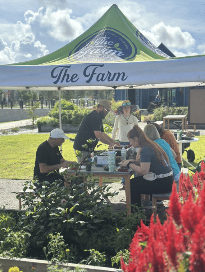 A group of people sits and stands around a table under a green and white tent labeled 'The Farm,' working together outdoors in a garden setting on a sunny day.